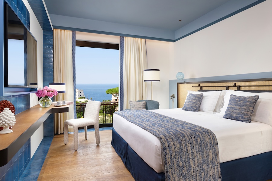 Hotel na Itália tem inspiração mediterrânea, paleta azul e vista para o mar. Na foto, quarto de casal, piso de madeira.
