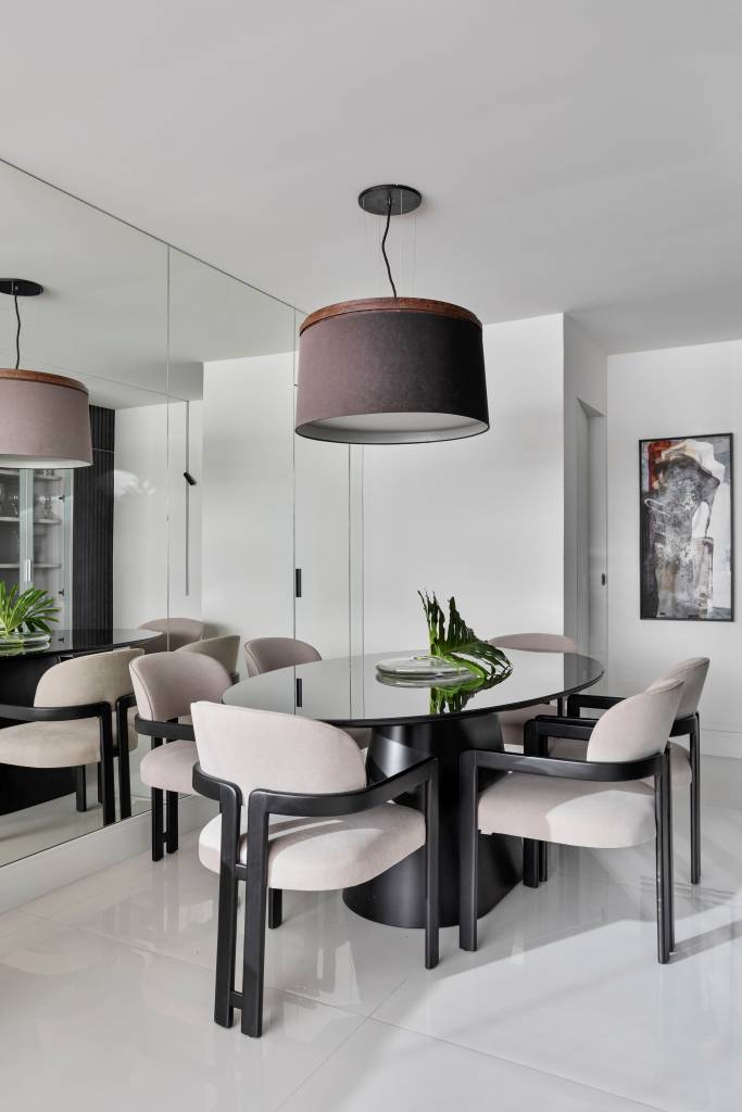 Com paleta ancorada no preto, apê tem painel ripado e piso de porcelanato. Projeto de Rebeca de França. Na foto, sala de jantar, espelho, mesa oval preta.
