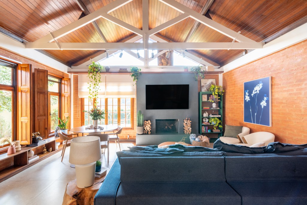 Sobrado de 160 m² em estilo rústico ganha cozinha verde com forno de pizza. Projeto de Abrazo Interiores. Na foto, teto de duas águas, sofá azul, lareira.