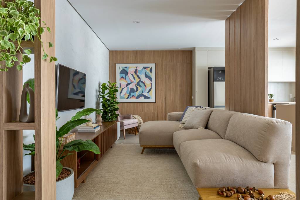 Apê ganha ilha de cozinha espaçosa e brises delimitando sala de TV. Projeto de Go Up Arquitetura. Na foto, sala, tv, sofá bege, tapete bege.