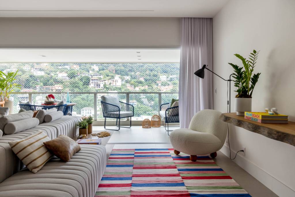 Azulejos pintados à mão com estampas botânicas revestem varanda de apê. Projeto de Renata Lemos. Na foto, sala de estar, tapete colorido listrado, poltrona branca.