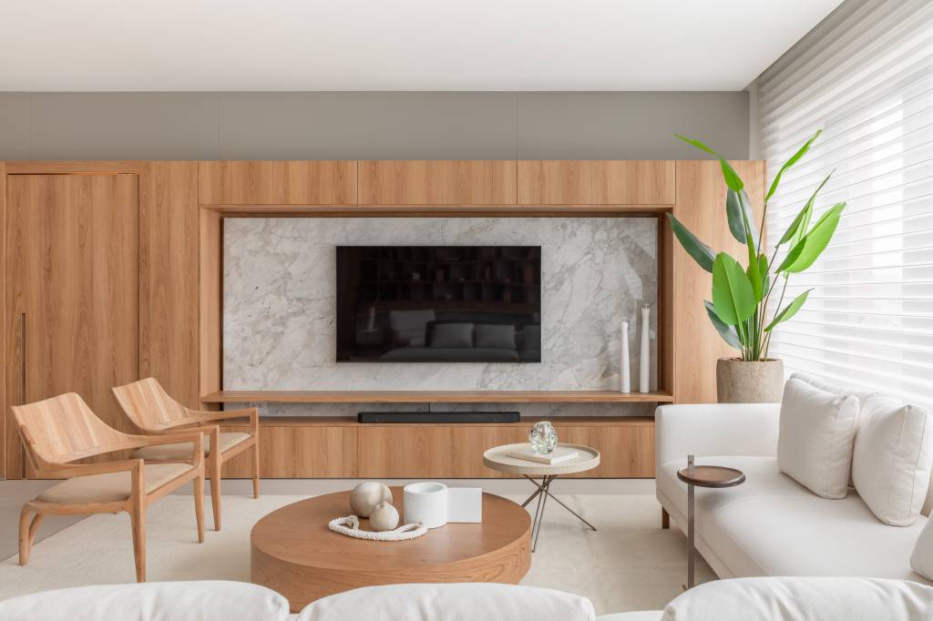 Biblioteca em sala de estar reflete paixão por leitura em apartamento. Projeto de NK Arquitetura. Na foto, sala de tv, mesa de centro redonda, planta, sofá branco.