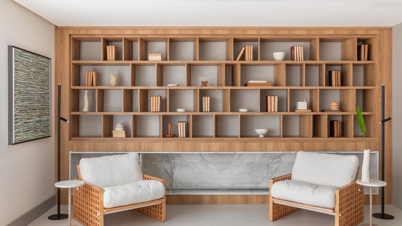 Biblioteca em sala de estar reflete paixão por leitura em apartamento. Projeto de NK Arquitetura. Na foto, sala de estar, estante de nichos com livros, poltrona branca.