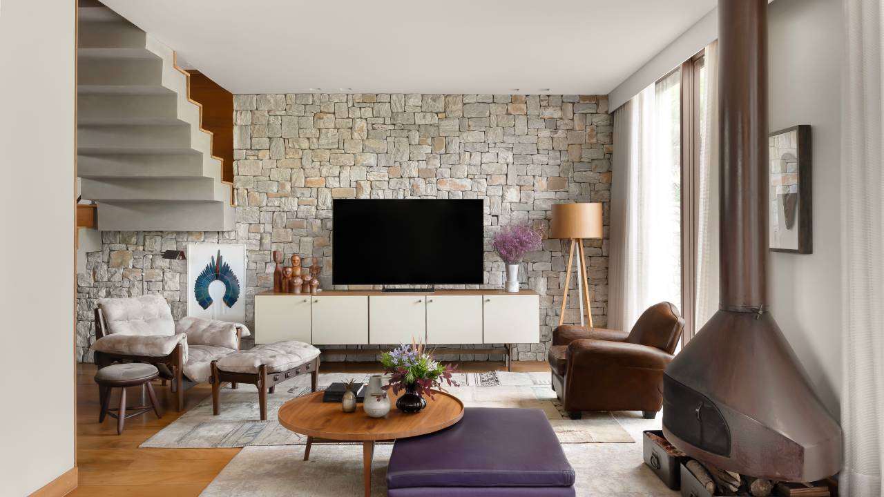 Sintonia com natureza: casa de campo com lareira tem sofá e cozinhas roxos. Projeto de Camila Medrado. Na foto, sala de estar, parede de pedras, pufe, mesa de centro.