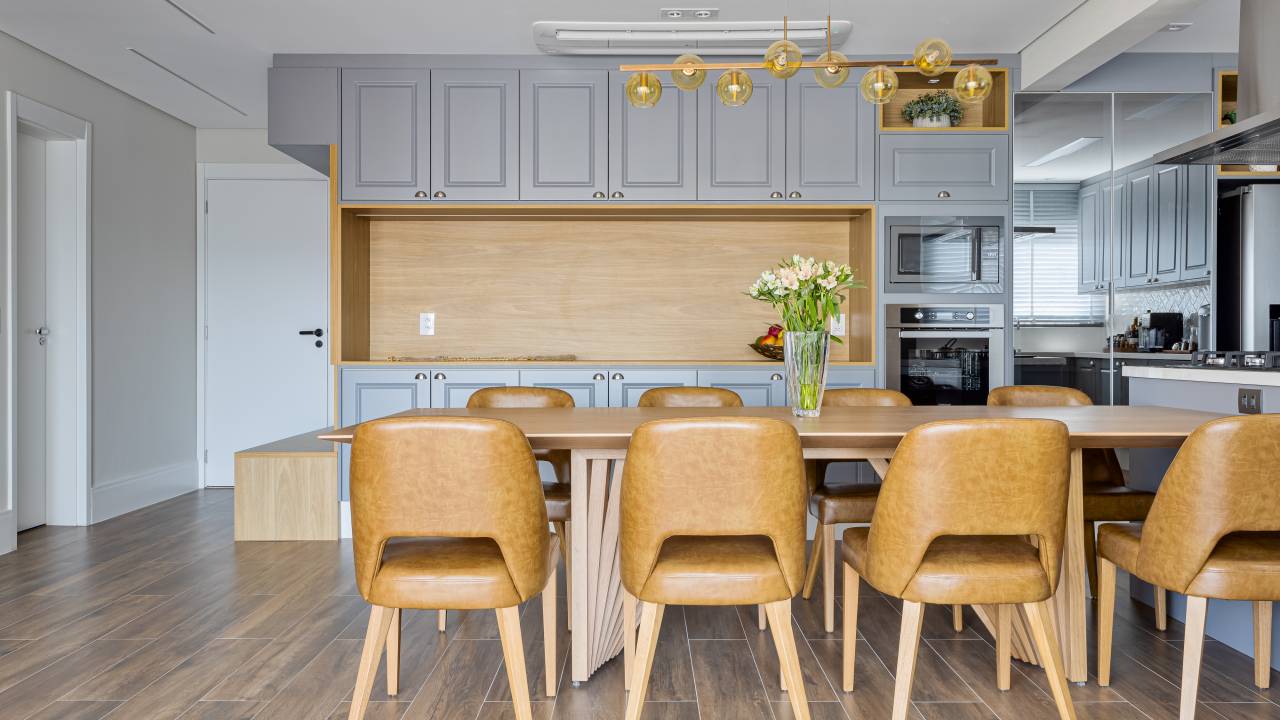 Cinza, branco, madeira e azul dão sensação de fluidez e tranquilidade neste apê. Projeto de Palladino Arquitetura. Na foto, cozinha integrada com jantar, cadeiras em couro caramelo, armário provençal.