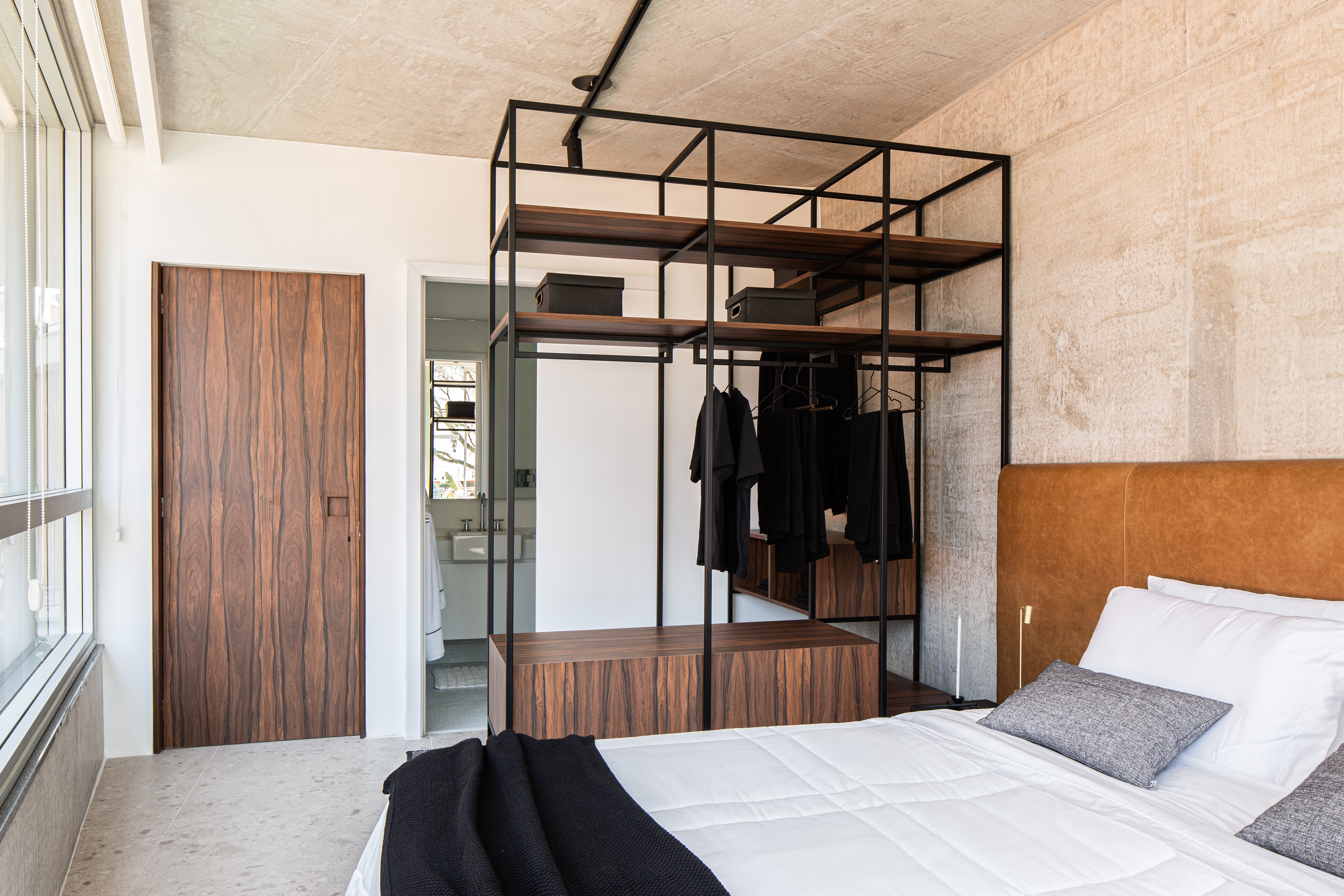Painéis em madeira pau ferro contrastam com laje de concreto neste apê. Projeto de Estúdio Pluri. Na foto, quarto, cama de casal, cabeceira de couro marrom, closet aberto.