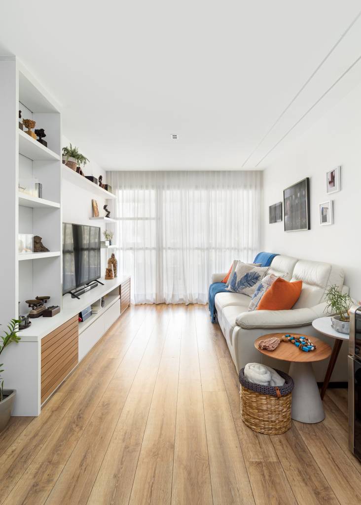 Parede entre sala e cozinha é demolida é dá lugar à cristaleira neste apê. Projeto de MA.DÊ Arquitetura. Na foto, sala de estar, sofá claro, estante branca com prateleiras e tv.