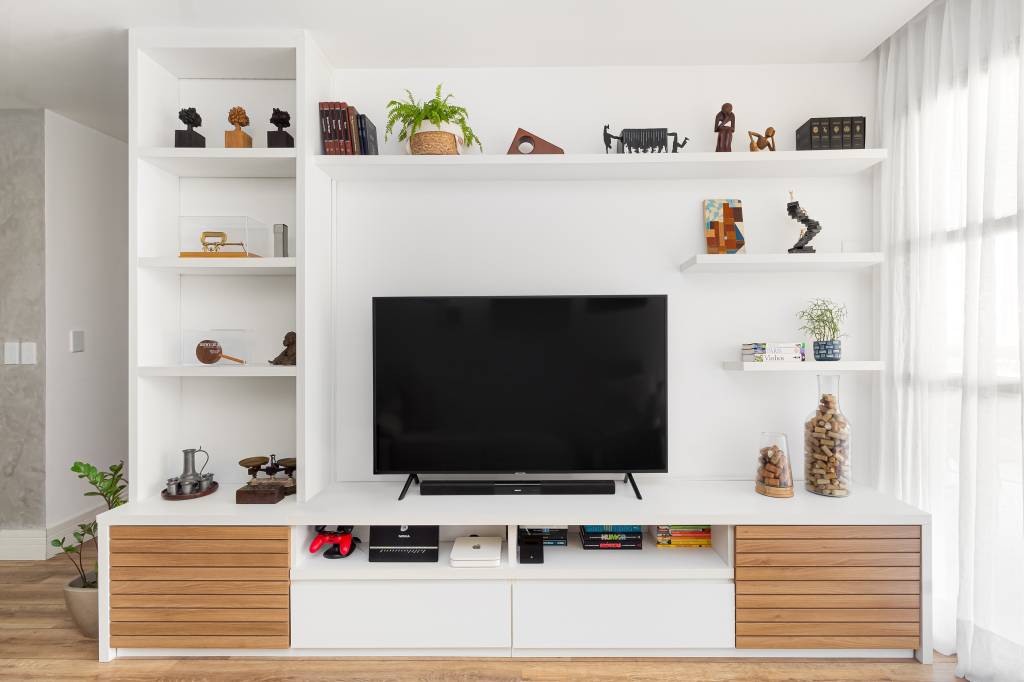 Parede entre sala e cozinha é demolida é dá lugar à cristaleira neste apê. Projeto de MA.DÊ Arquitetura. Na foto, sala de estar, sofá claro, estante branca com prateleiras e tv.