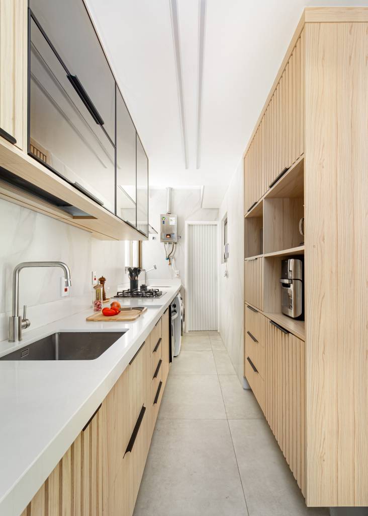 Parede entre sala e cozinha é demolida é dá lugar à cristaleira neste apê. Projeto de MA.DÊ Arquitetura. Na foto, cozinha, corredor, armário em serralheria e ripado, bancada branca.