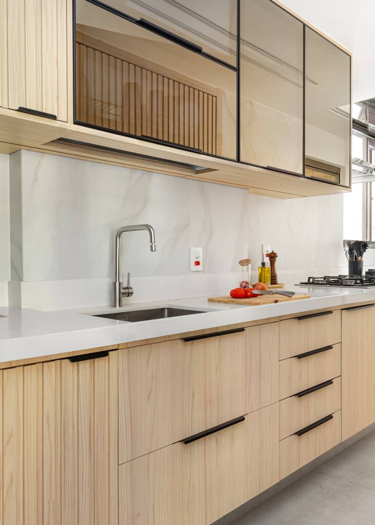 Parede entre sala e cozinha é demolida é dá lugar à cristaleira neste apê. Projeto de MA.DÊ Arquitetura. Na foto, cozinha, corredor, armário em serralheria e ripado, bancada branca.