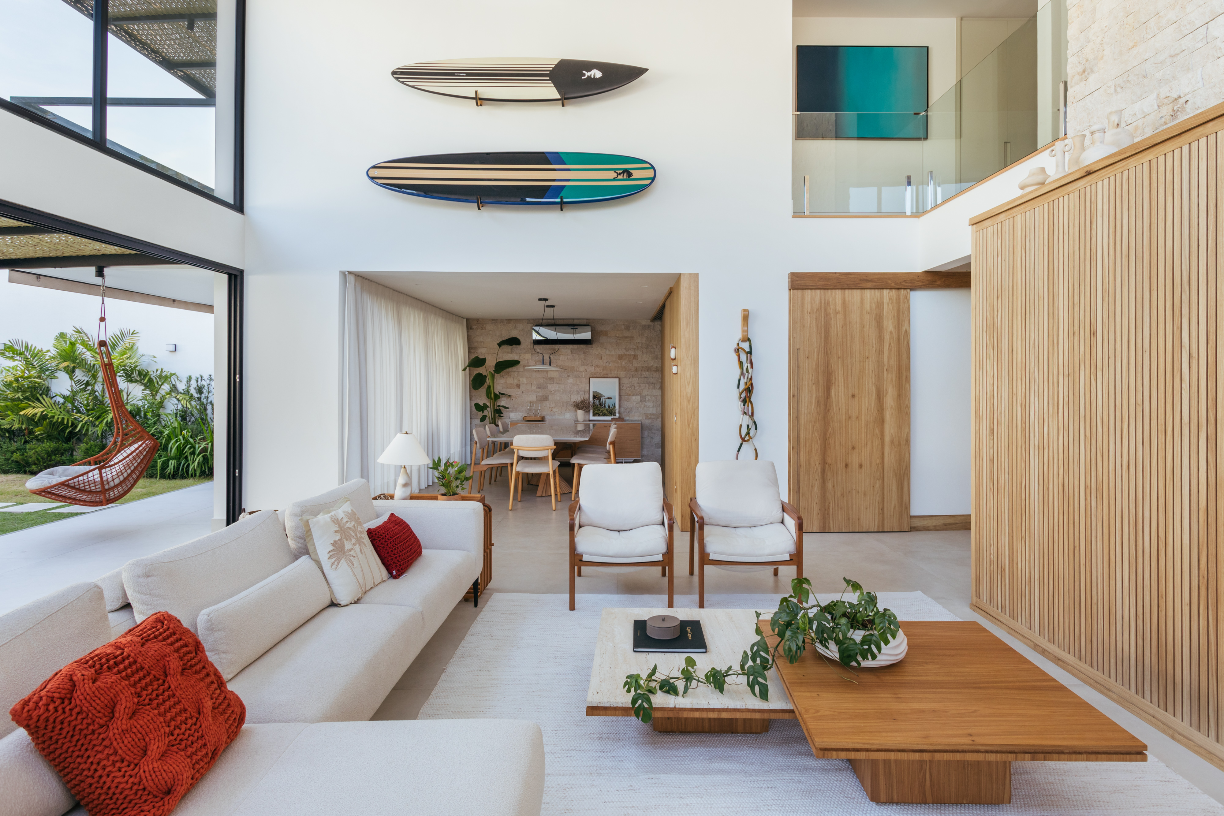 Pergolados com forro em fibras de bambu trançadas envolvem casa de 195 m². Projeto de Amanda Miranda. Na foto, sala de estar com pé-direito duplo, pranchas de surfe na parede.
