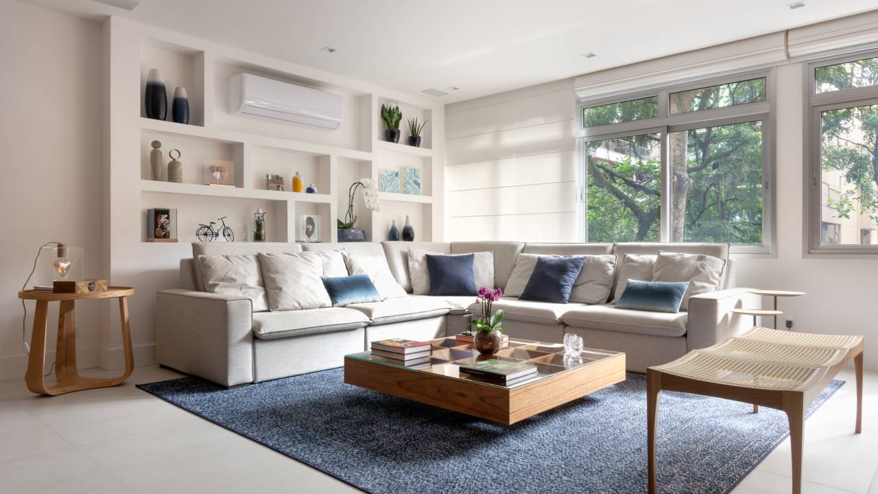 Reforma cria estante em gesso acartonado para acomodar tubulações. Projeto de Fernanda Medeiros. Na foto, sala, estante de nichos, ar condicionado, tapete azul, sofá branco.
