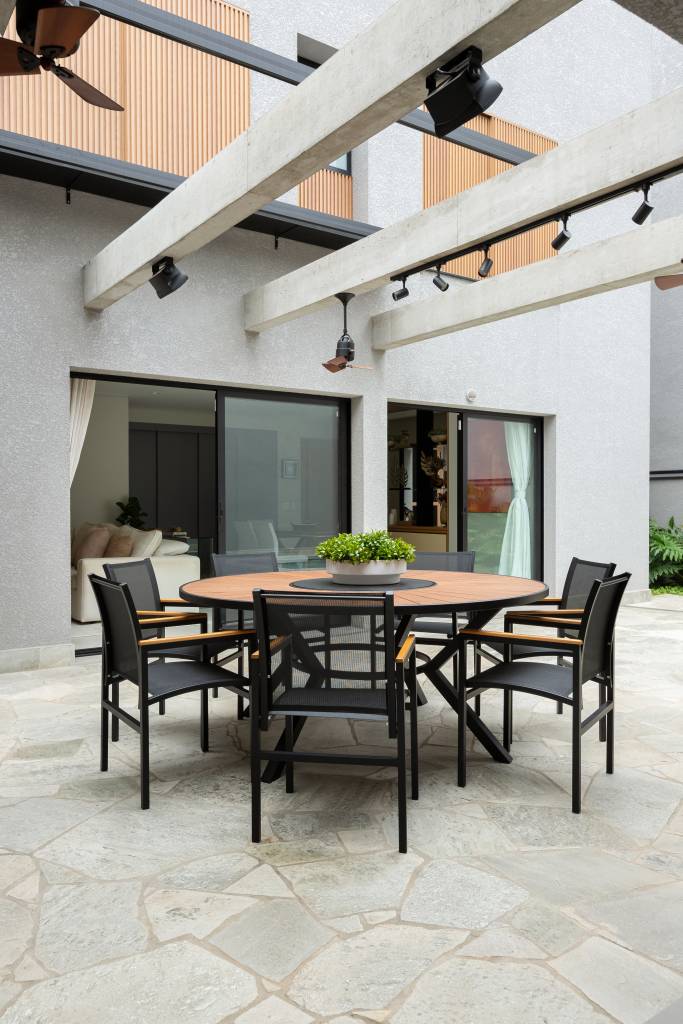 Casa tem área gourmet com toldo retrátil e piso em pedra rústica. Projeto de Gabriela Toledo Arquitetura. Na foto, mesa redonda, cadeiras pretas.