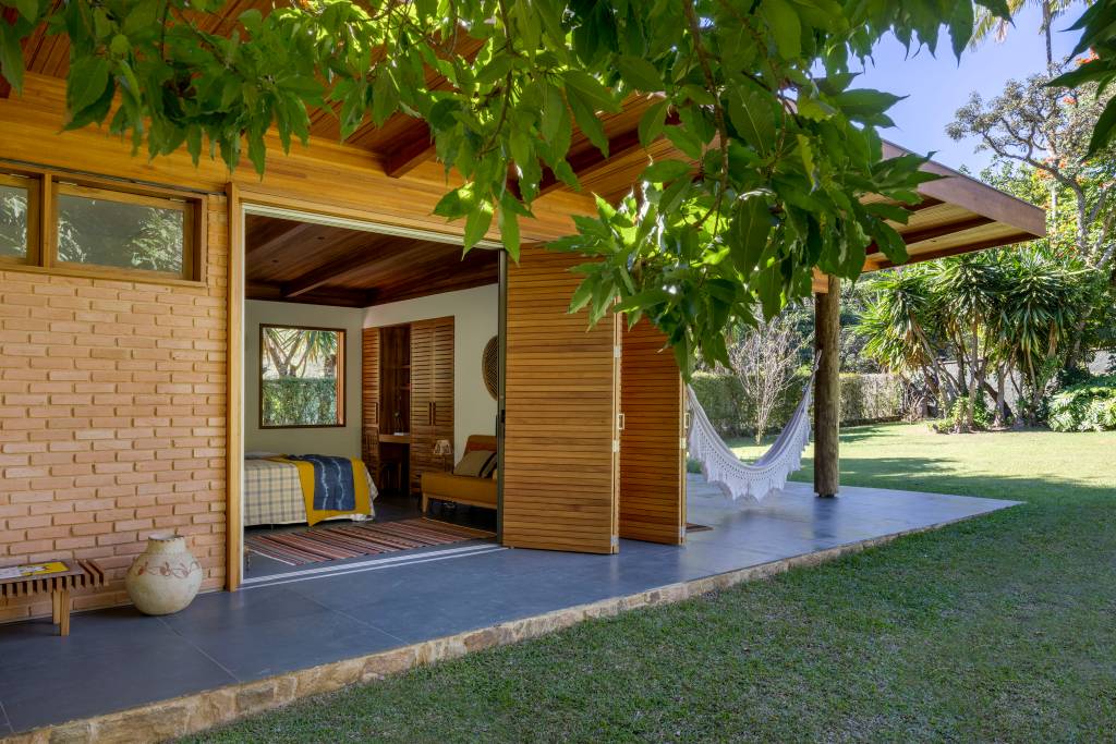 Casa de campo de hóspedes tem teto com madeira de demolição e piso de pedra ardósia. Projeto de Fabiana Cyon e Ana Paula Castro. Na foto, varanda, árvore, porta camarão.