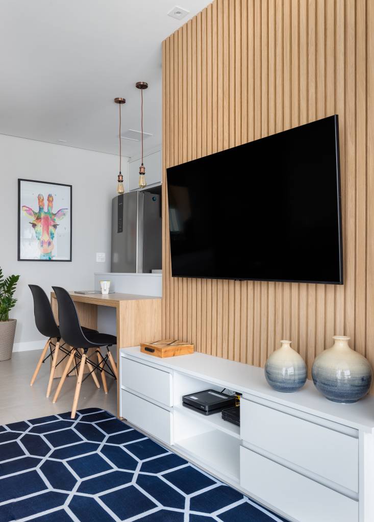 Cozinha com gabinete azul e painel ripado na sala dão textura e cor a apê de 62 m². Projeto de Duno Arquitetura. Na foto, sala de tv, tapete azul, rack branco.