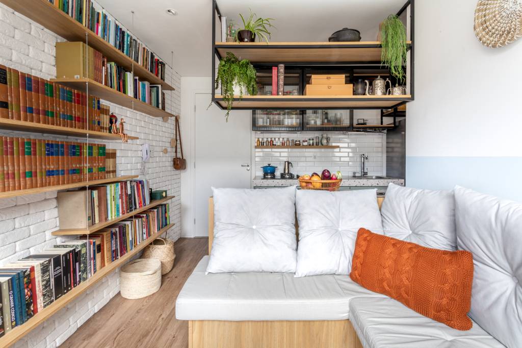 Miniapê de 25 m² tem sofá de canto alemão com baú e estante de mil livros. Projeto de Lemarchi Arquitetura. Na foto, cozinha integrada, prateleira com livros.