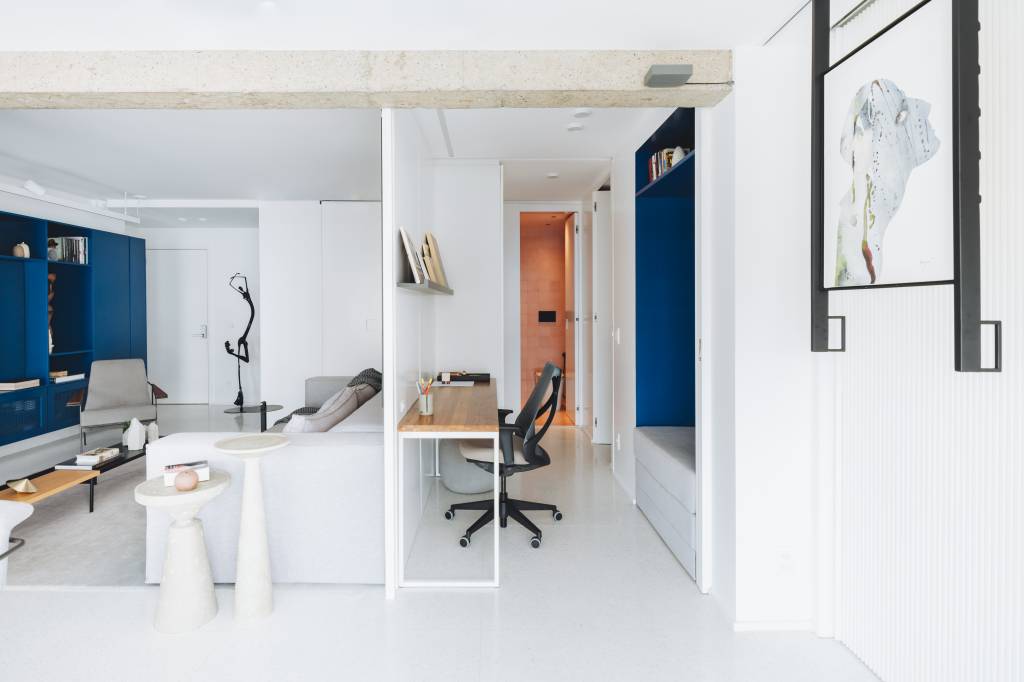 Piso de granilite e revestimentos claros são pano de fundo para arte neste apê. Projeto de Projeto Rawi Arquitetura + Design. Na foto, sala de estar integrada com home office, painel de marcenaria branco.