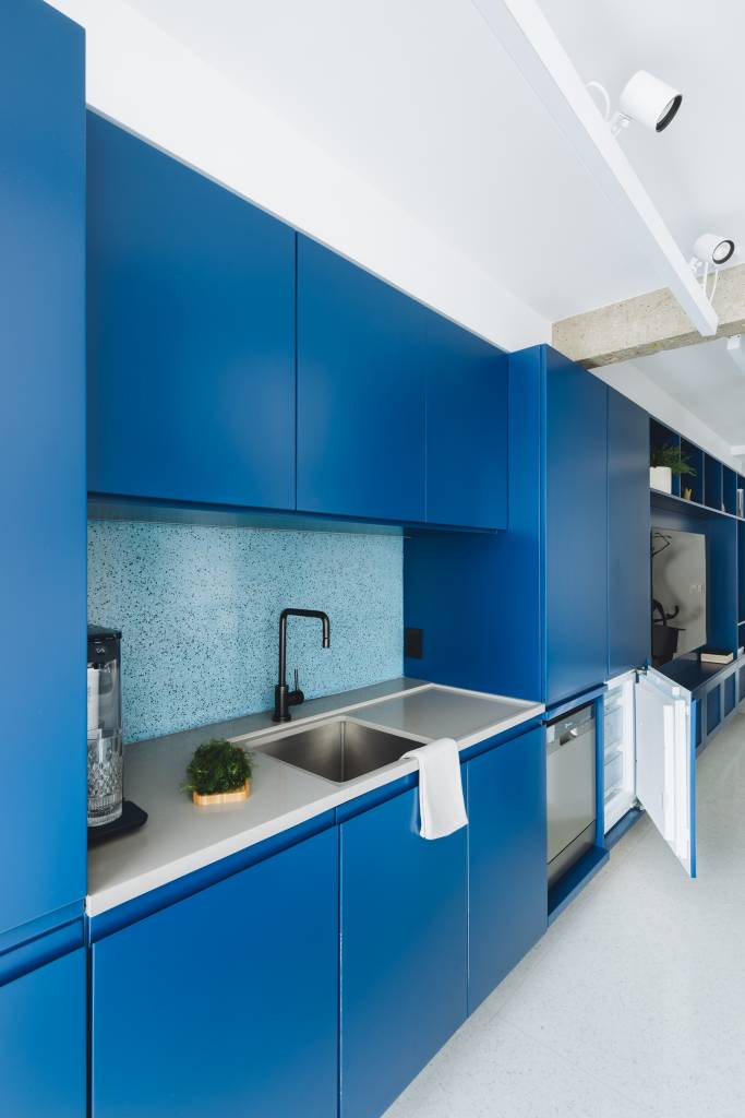 Piso de granilite e revestimentos claros são pano de fundo para arte neste apê. Projeto de Projeto Rawi Arquitetura + Design. Na foto, cozinha, marcenaria azul, backsplash de azulejos azuis.