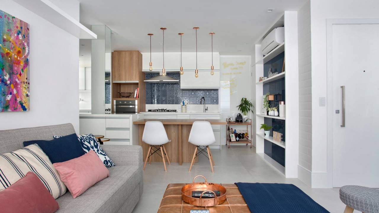 Estante com fundo de vidro é criada com integração entre sala e cozinha de apê de 68 m². Projeto da designer de interiores Claudia Infante. Na foto, sofá cinza, dois quadros acima do sofá, pufe de couro como mesa de centro, banco de marcenaria, bancada de refeições.