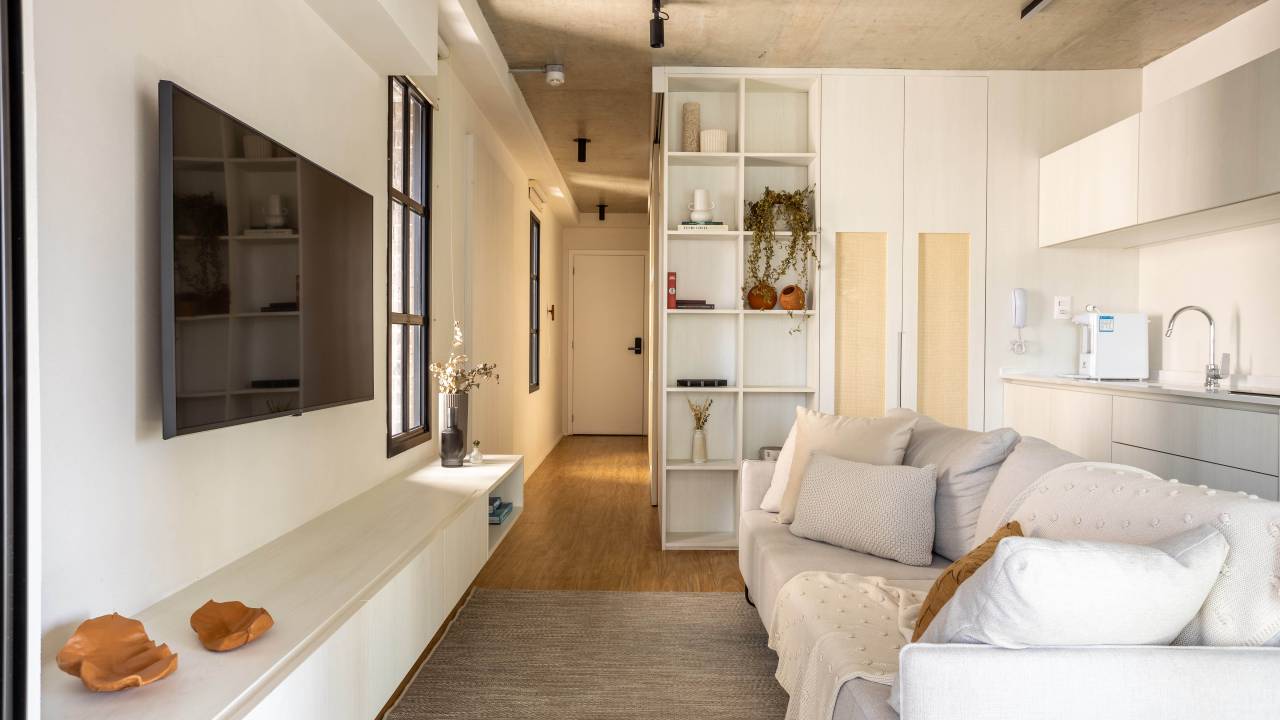 Loft de 48 m² segue estilo wabi sabi e tem palhinha nas portas de correr. Projeto de Carolina Gava. Na foto, sala de estar pequena, sofá branco, rack baixo branco.