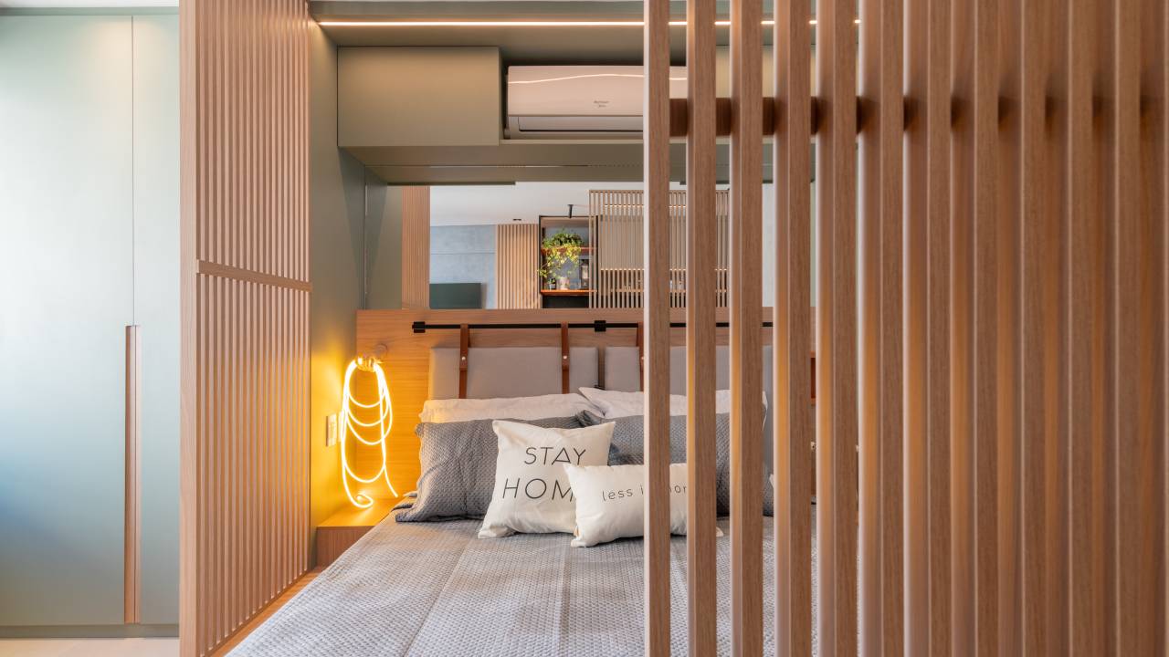 Painéis ripados vazados emolduram cama em estúdio de apenas 26 m². Projeto de Carolina Bordonco Interiores. Na foto, quarto, luminária, ar condicionado.