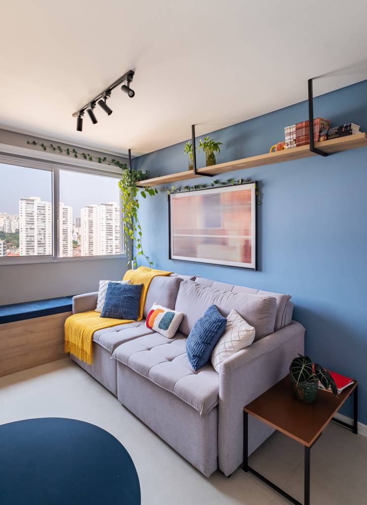 Projeto de iluminação ressalta o azul neste apartamento de 34 m². Projeto Dua Arquitetura e Bya Ribeiro. Na foto, sala, sofá cinza, quadro, prateleira.