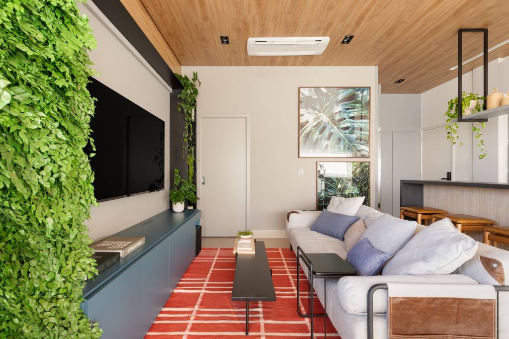 Apê de 100 m² com pé-direto de 3 m ganha soluções criativas. Projeto de Michelle Machado. Na foto, sala de estar com tapete vermelho, jardim vertical e TV.