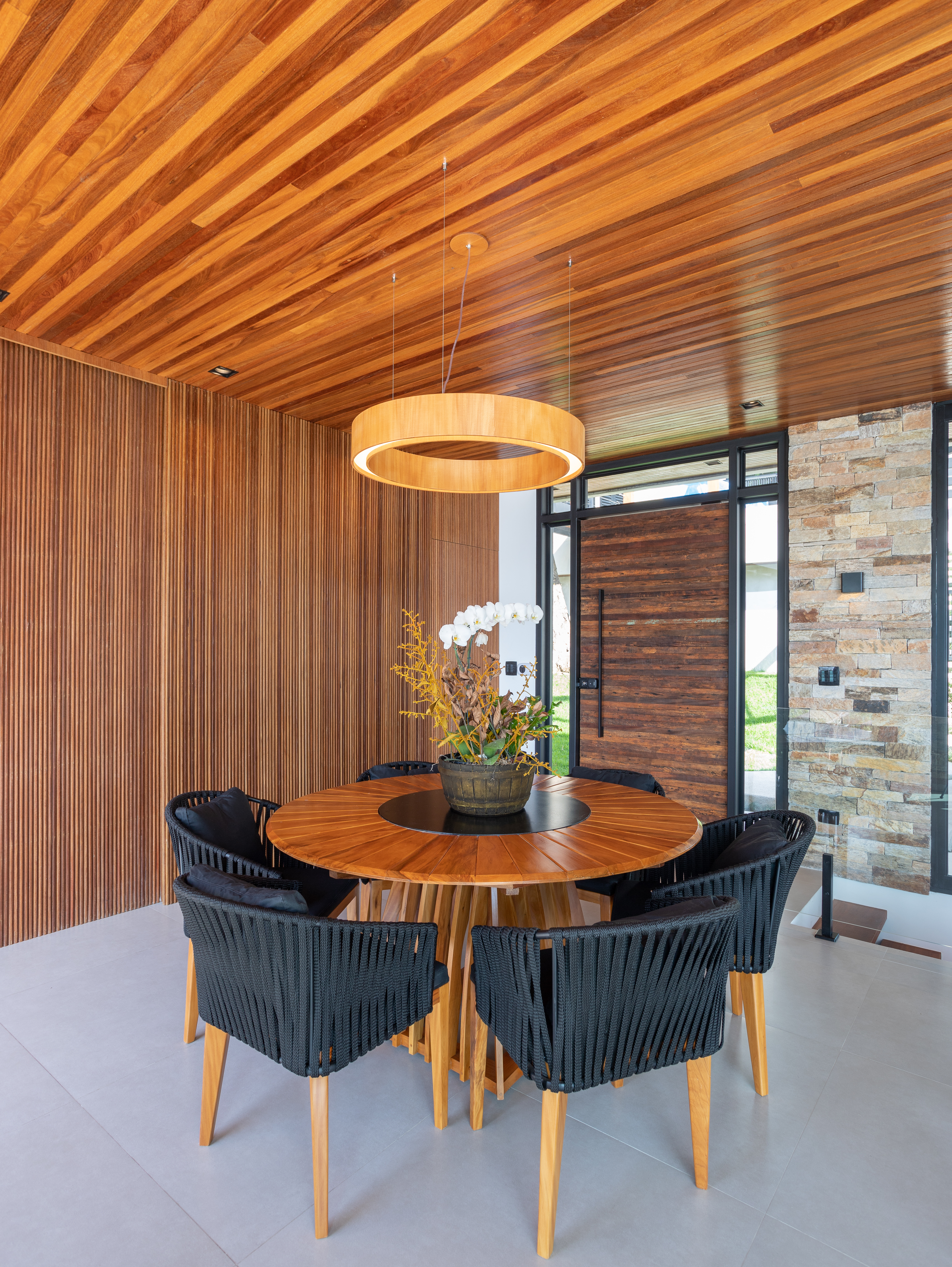 Casa de praia com varanda tem forro e painel ripado para criar estética elegante. Projeto de Ana Gadioli Arquitetura. Na foto, mesa de madeira redonda, cadeiras pretas estofadas.