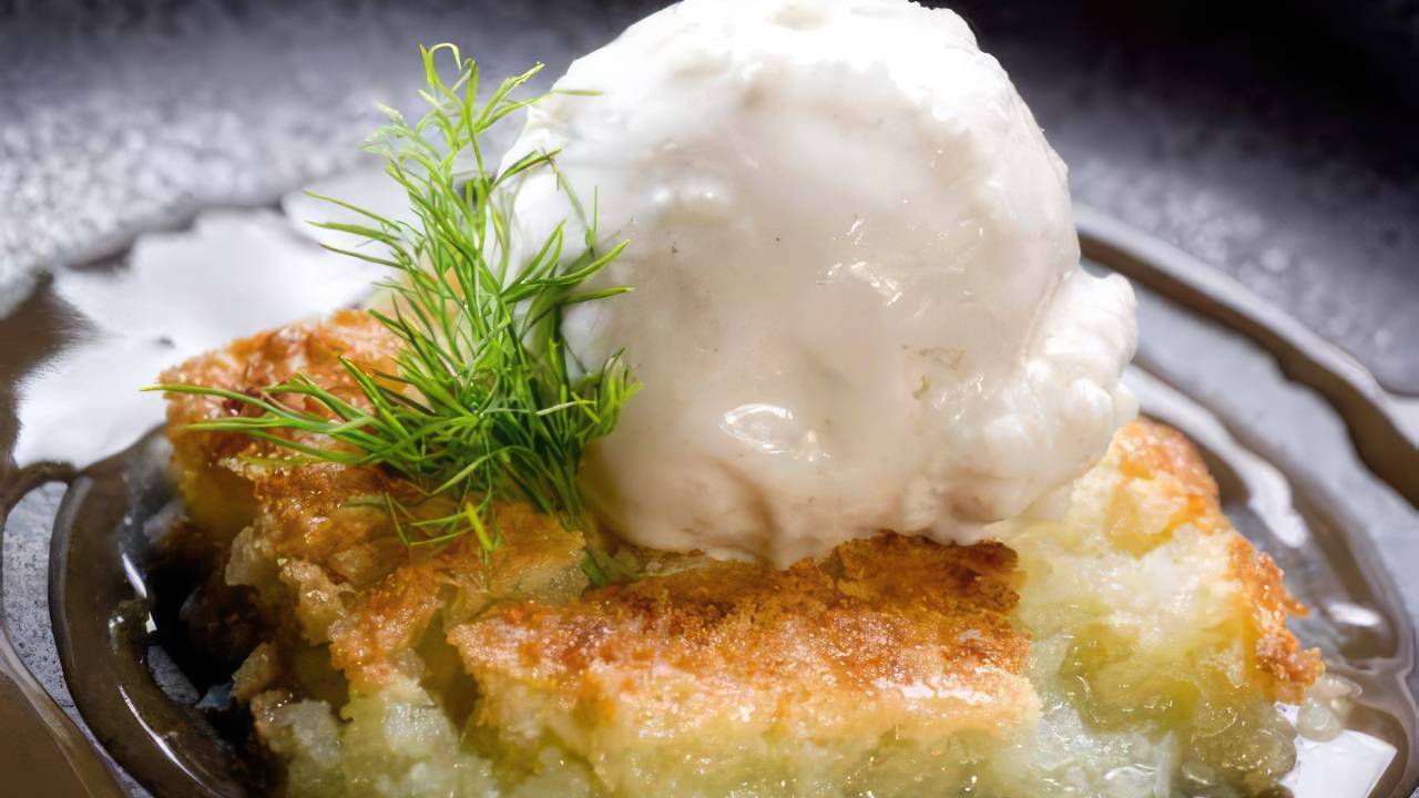 A imagem mostra uma porção generosa de cocada de tabuleiro, uma sobremesa tradicional brasileira feita com coco ralado, açúcar e ovos, disposta elegantemente em um prato. Ao lado, uma bola de sorvete de tapioca, que se derrete levemente ao contato com a cocada quente, formando uma combinação perfeita de texturas e sabores. O fundo é simples, permitindo que o foco permaneça na deliciosa sobremesa.