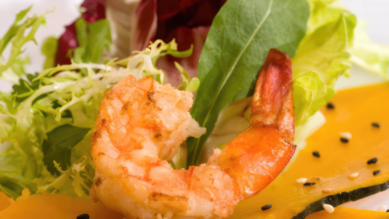 Imagem de uma salada de camarão imperial, destacando lâminas de abóbora cabotiã cozida em leite de coco, cobertas com camarões grelhados e polvilhadas com gergelim. A salada é disposta em um prato elegante, rodeada por folhas verdes frescas, apresentando uma combinação vibrante de cores e texturas que valorizam a gastronomia brasileira.