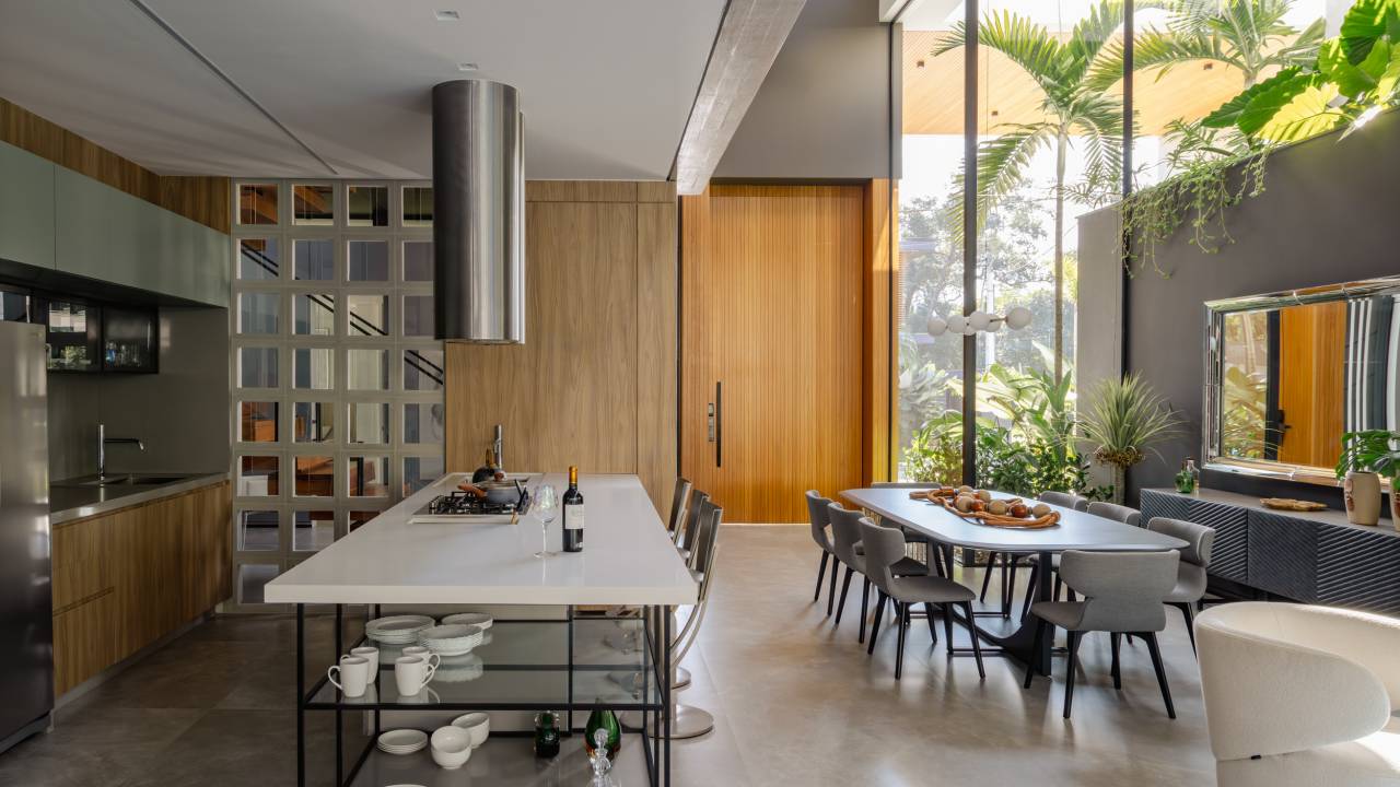 Casa possui áreas sociais envolvidas por vidro e vista para a Mata Atlântica. Projeto de Raiz Arquitetura. Na foto, cozinha com ilha central e mesa de jantar na lateral.