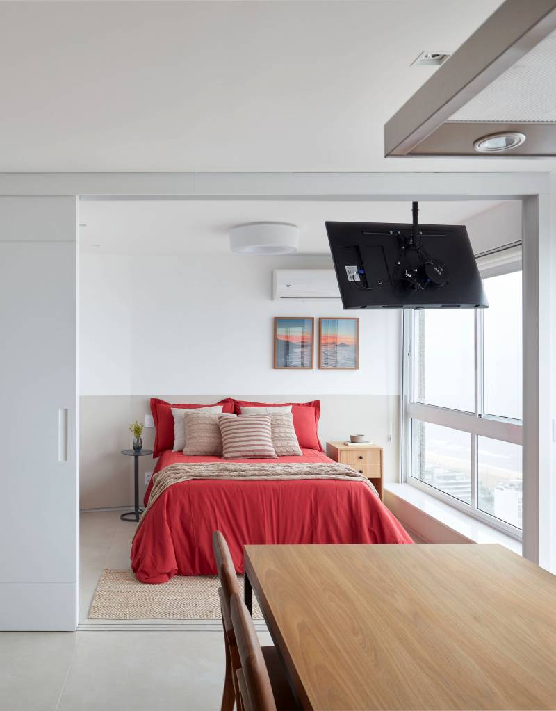 Apê de 50 m² tem portas de correr e bancada de trabalho na estante. Projeto de Carolina Gouvea Arquitetura. Na foto, quarto, roupa de cama vermelha.