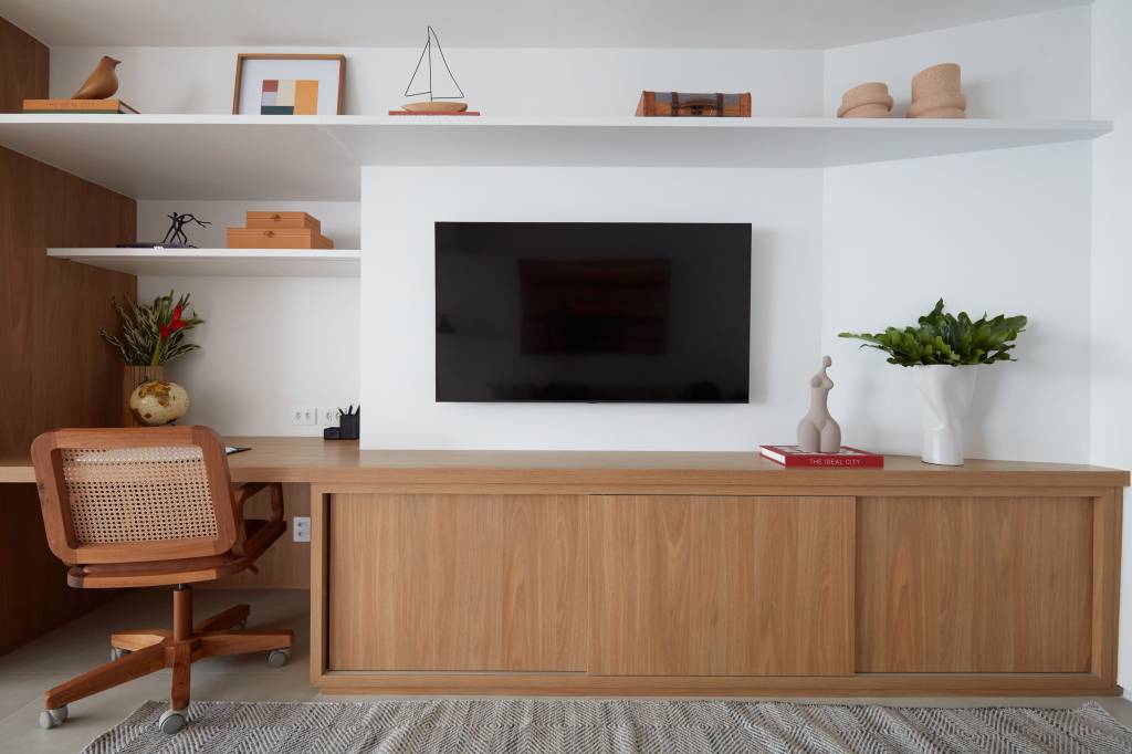 Apê de 50 m² tem portas de correr e bancada de trabalho na estante. Projeto de Carolina Gouvea Arquitetura. Na foto, sala, tv, home office pequeno.