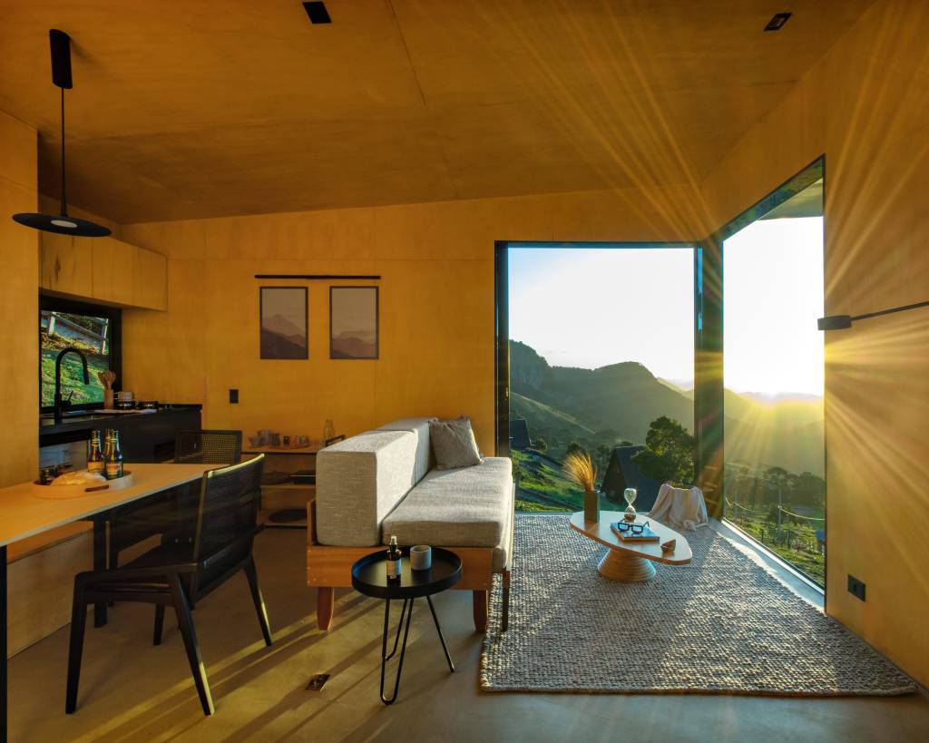 Chalé de apenas 48 m² tem paredes com painéis de madeira e vista para serra. Projeto de RAWI Arquitetura+Design. Na foto, sala, sofá cinza, grandes janelas.