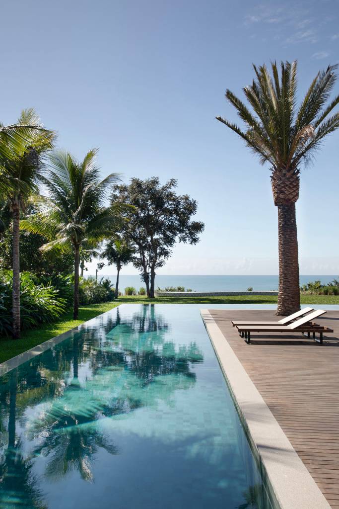 Teto de casa de praia tem forro em madeira cumaru com palha de dendê trançada. Projeto de David Bastos. Na foto, jardim, palmeiras, piscina em formato L.
