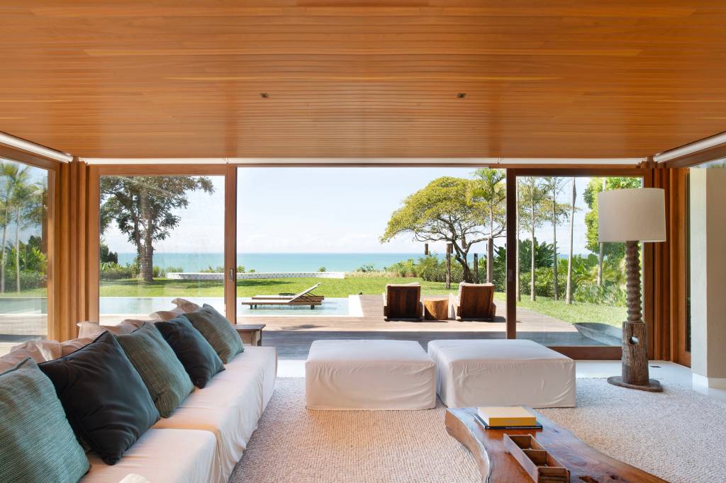 Teto de casa de praia tem forro em madeira cumaru com palha de dendê trançada. Projeto de David Bastos. Na foto, sala, sofá branco, vista para piscina.