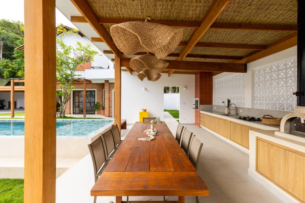 Casa com clima de praia em São José dos Campos. Projeto de escritório Sertão Arquitetos. Na foto, área da churrasqueira com mesa extensa.