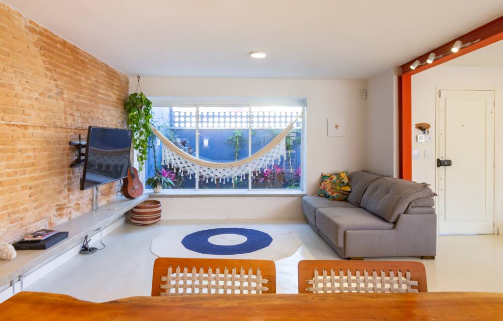 Casa com parede de tijolinhos e detalhes em cores primárias. Projeto de Sertão Arquitetos. Na foto, sala de estar com rede perto da janela.