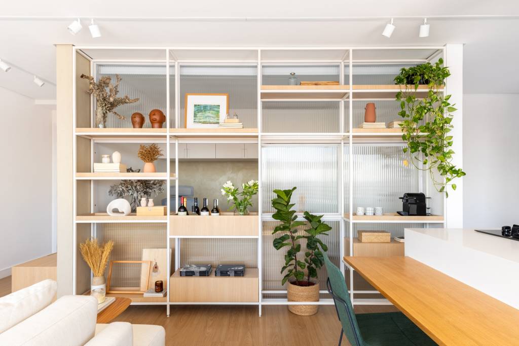 Estante de marcenaria, vidro e serralheria é divisória entre home office e sala neste apê 