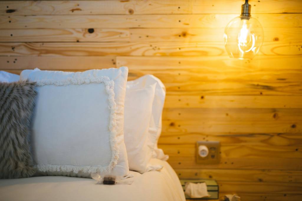 cama-de-inverno-6-dicas-para-deixar-seu-quarto-aconchegante-e-quentinho_2 Detalhe de uma cama com diversos travesseiros. Ao fundo, uma parede de madeira.