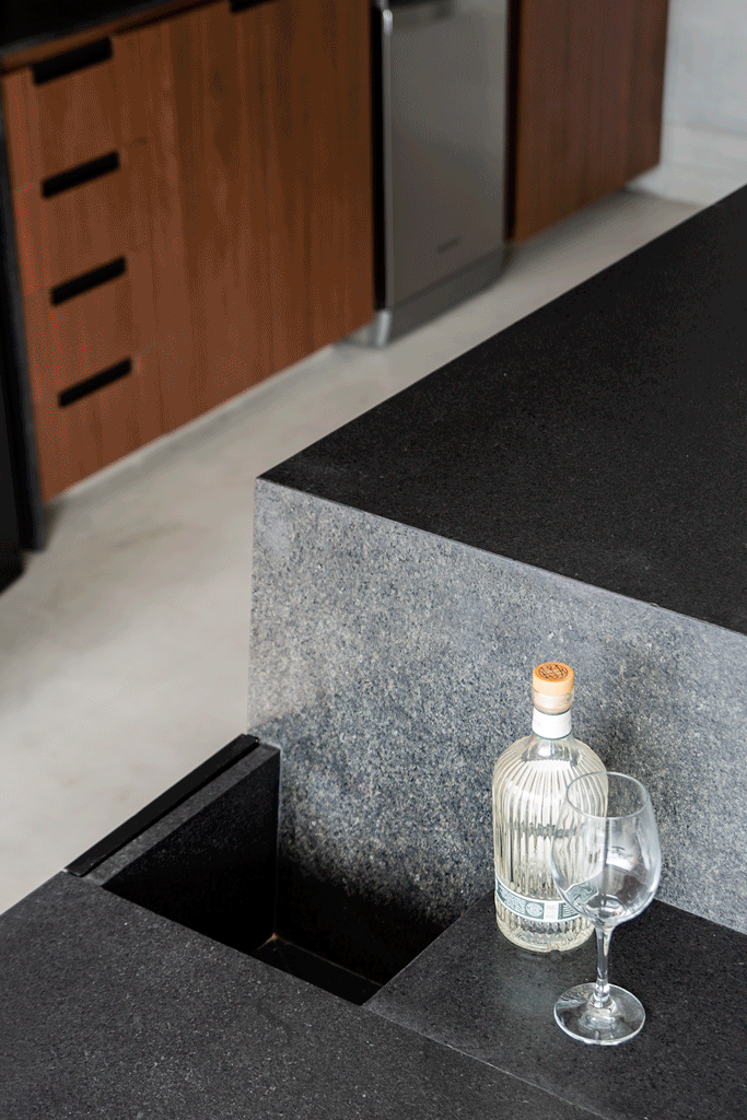 Apê industrial tem champanheira embutida na cozinha e porcelanato na parede do quarto. Projeto de Zalc Arquitetura. Na foto, Bancada de cozinha moderna em granito preto com pia integrada, garrafa de gin e taça de vinho vazia. Ao fundo, armários de madeira e eletrodoméstico de aço inoxidável