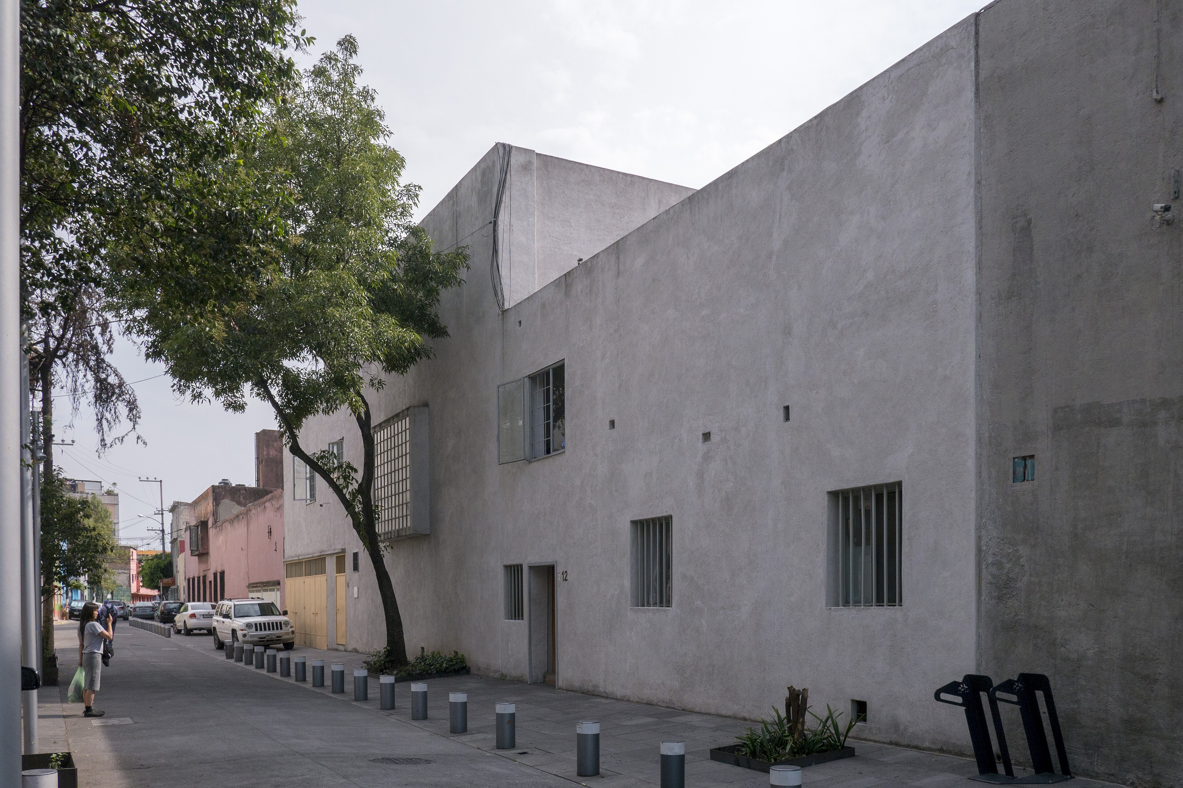 Rua com calçada de concreto e postes cinzas, um prédio cinza claro com janelas gradeadas e uma árvore frondosa. À esquerda, uma pessoa de camiseta clara e calça escura observa carros estacionados.