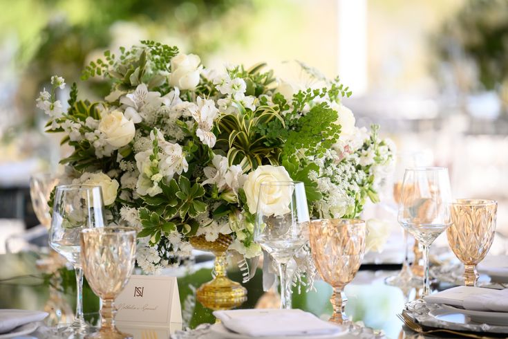 Mesa posta para evento com arranjo floral central de rosas brancas e folhagens verdes, taças de cristal transparentes e âmbar, e um cartão de lugar com as iniciais "NS"