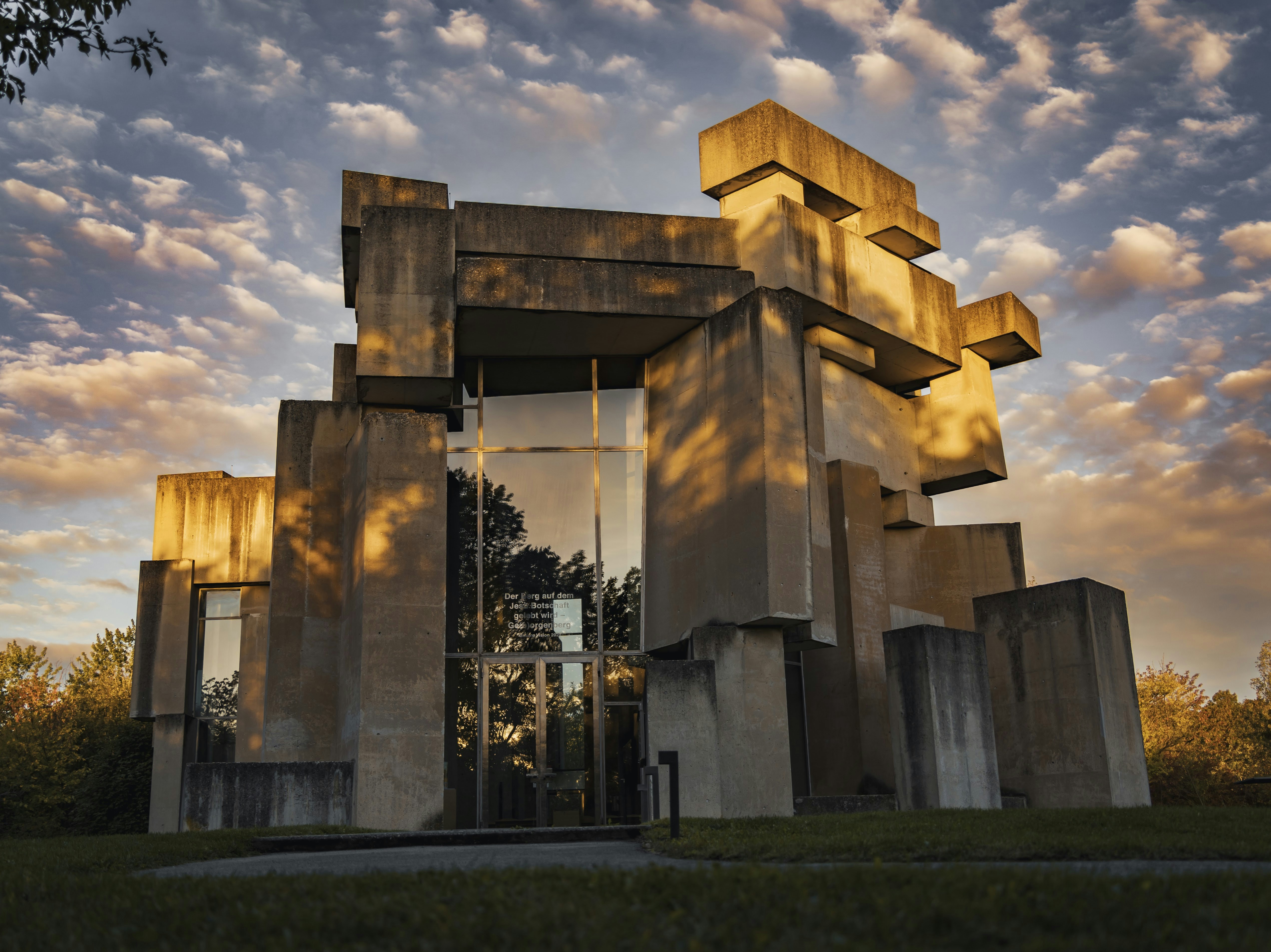 Edifício brutalista de concreto com blocos empilhados e janelas de vidro refletindo árvores e o céu. A luz dourada do pôr do sol ilumina a fachada, criando sombras dramáticas. O céu tem nuvens esparsas e tons alaranjados e azuis. Há grama verde na frente e árvores ao redor