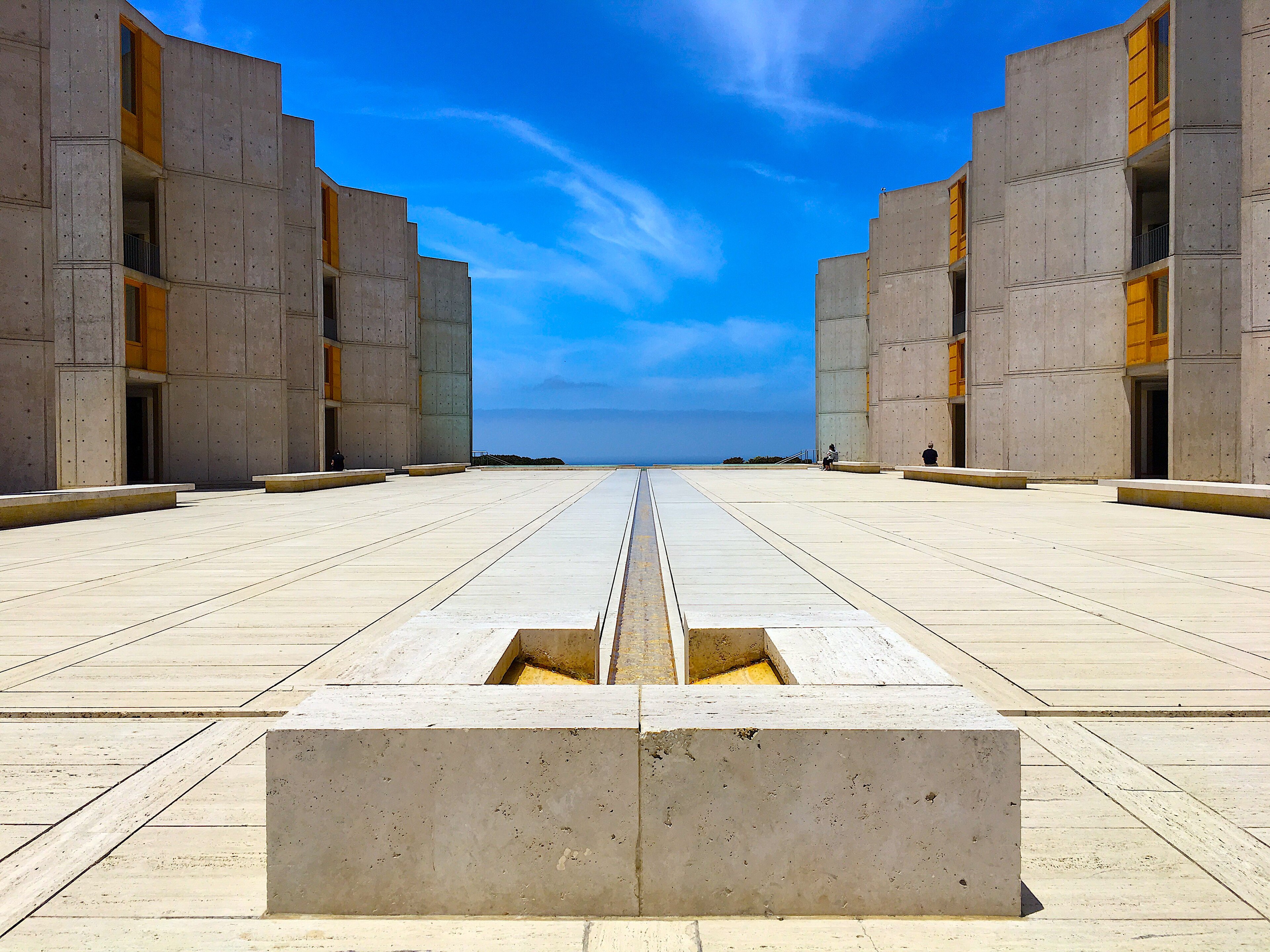 Pátio amplo com piso de pedra clara, ladeado por dois edifícios de concreto aparente com janelas de madeira amarela. Ao fundo, o oceano sob um céu azul vibrante com nuvens esparsas. Duas pessoas sentadas em bancos de pedra, distantes. Um canal estreito de água divide o pátio centralmente.