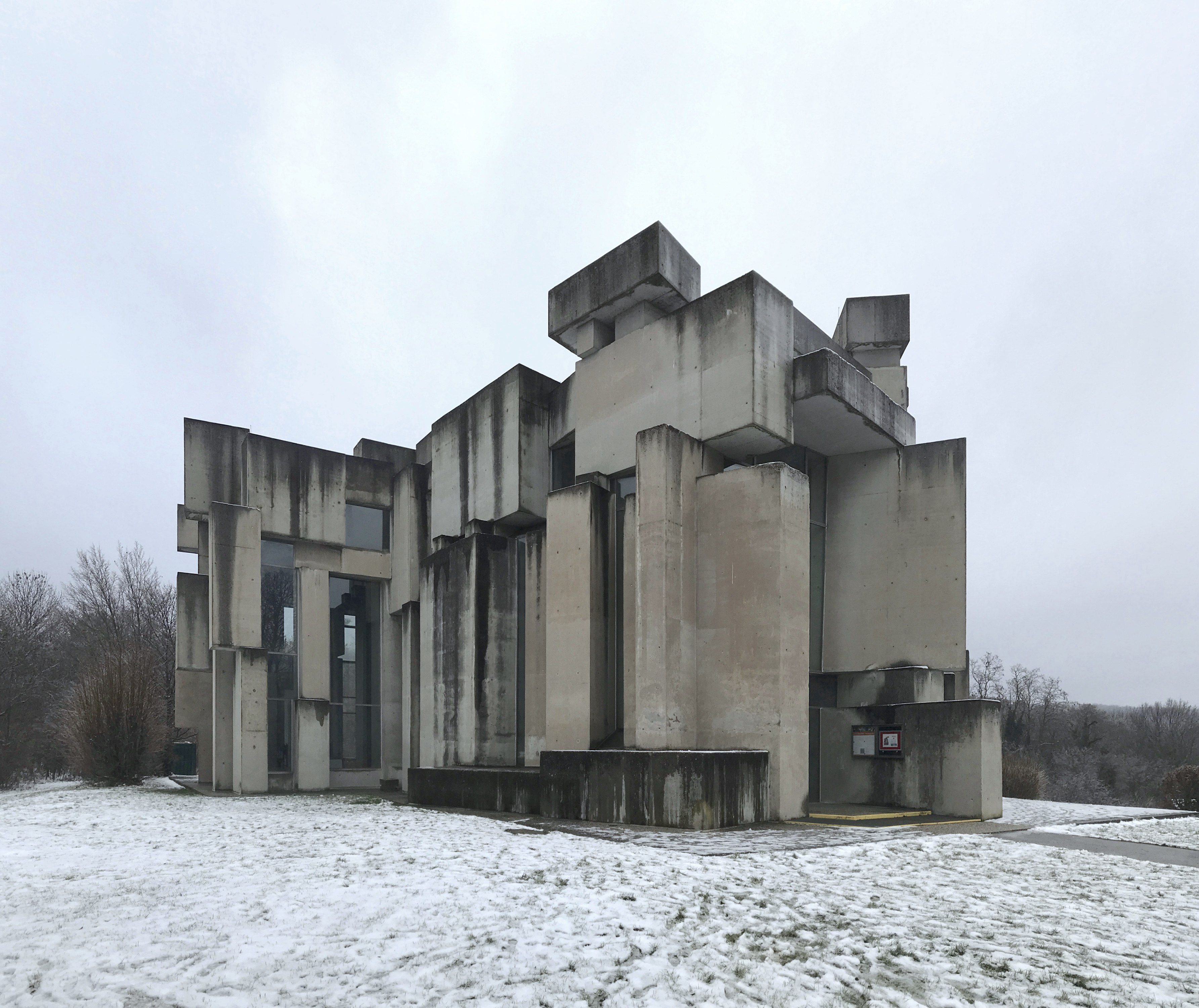 Edifício brutalista de concreto cinza com blocos e pilares irregulares, janelas escuras, em terreno coberto por neve e céu nublado