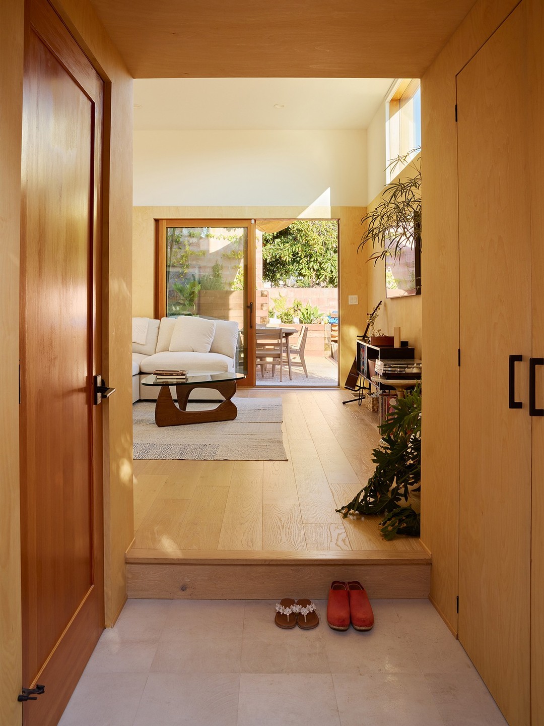 Entrada de uma casa moderna com porta de madeira à esquerda, piso de tábuas claras e um degrau. À frente, sala com sofá branco, mesa de centro e porta de vidro que dá para um pátio externo com mesa e cadeiras. À direita, armários de madeira clara e plantas. Dois pares de chinelos estão no chão, perto do degrau