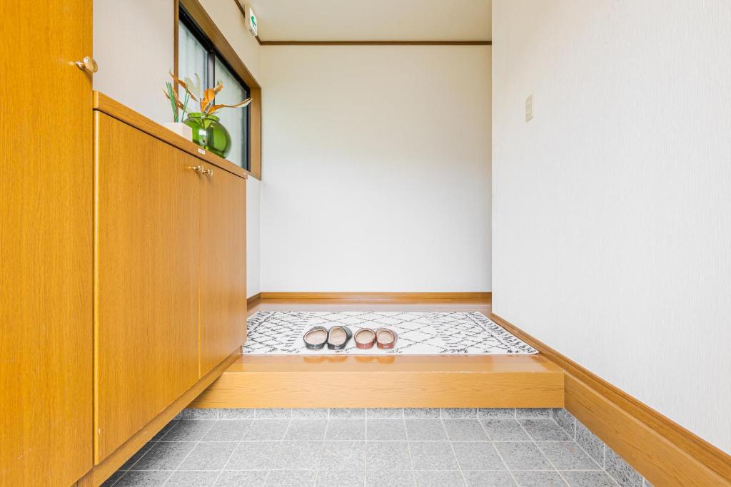 Entrada de uma casa com piso de cerâmica cinza, um degrau de madeira clara, e um tapete branco com desenhos geométricos pretos. Dois pares de chinelos estão sobre o tapete. À esquerda, um armário de madeira clara com um vaso de plantas e uma janela acima. As paredes são brancas