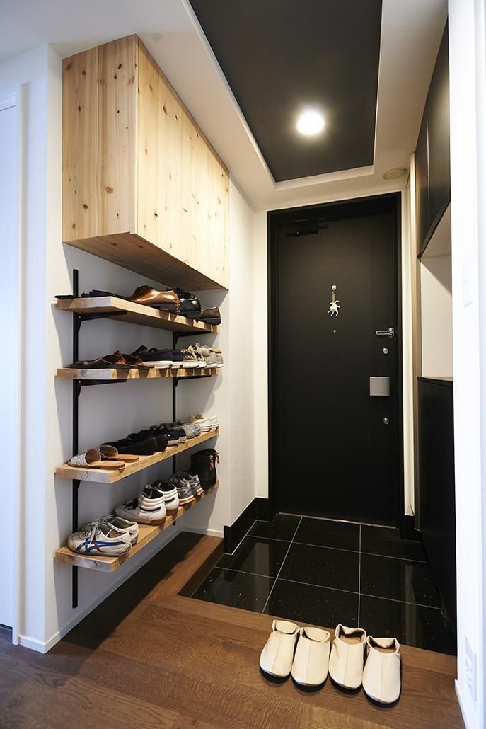 Entrada de casa com sapateira de madeira clara e prateleiras pretas cheias de sapatos, porta preta com chaveiro de caranguejo e piso de madeira e azulejo preto. Quatro pares de chinelos brancos estão no chão