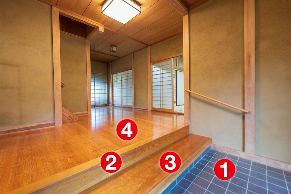 Entrada de uma casa japonesa com piso de cerâmica escura, dois degraus de madeira clara e um corrimão simples. Ao fundo, piso de madeira brilhante e portas de correr shoji com treliças de madeira e papel translúcido, iluminadas por luz natural e luminárias retangulares no teto de madeira